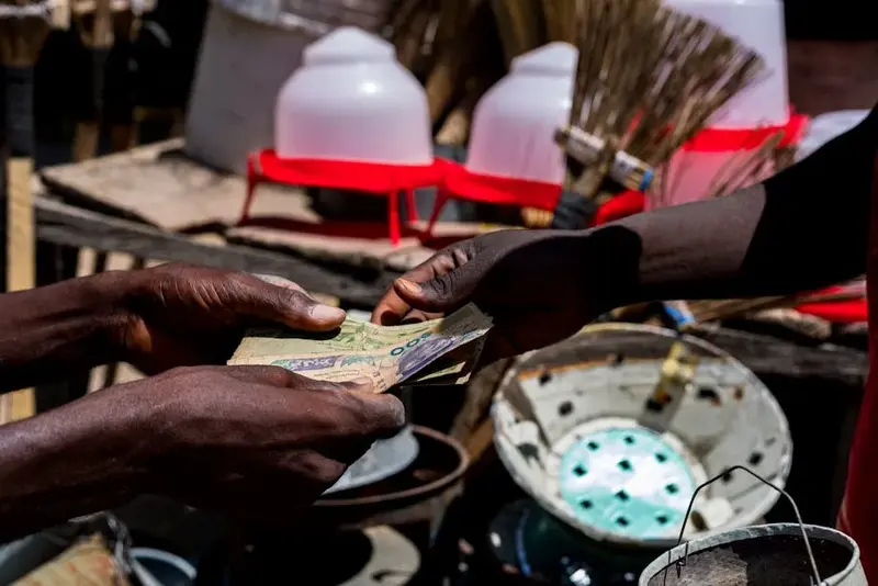 Close-up of hands exchanging Nigerian naira bills at a bustling outdoor market in Nigeria.